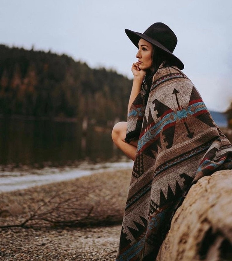 May include: A woman wearing a black hat sits on a log by a lake, wrapped in a brown, blue, and red patterned blanket. The blanket has a geometric design with arrows and other symbols.
