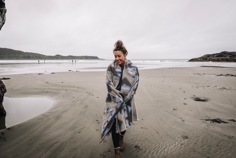 May include: A woman walks on a sandy beach wearing a blue and brown patterned blanket. The beach is empty except for a few people in the distance.