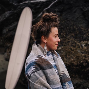 May include: A woman with brown hair pulled back in a bun is wearing a grey and blue patterned blanket. She is standing in front of a rocky background and looking to the right.
