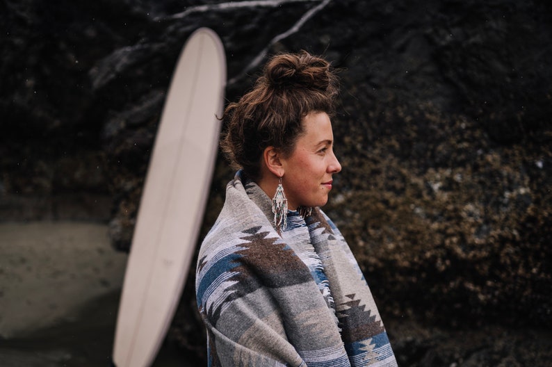 May include: A woman with brown hair pulled back in a bun is wearing a gray and blue patterned blanket. She is standing in front of a rocky background and looking to the right.