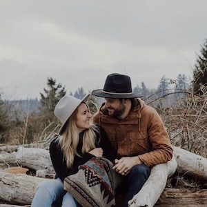 May include: A couple wearing hats and a blanket sit on a log in a wooded area. The woman is wearing a gray hat and a black sweater. The man is wearing a brown jacket and a black hat.