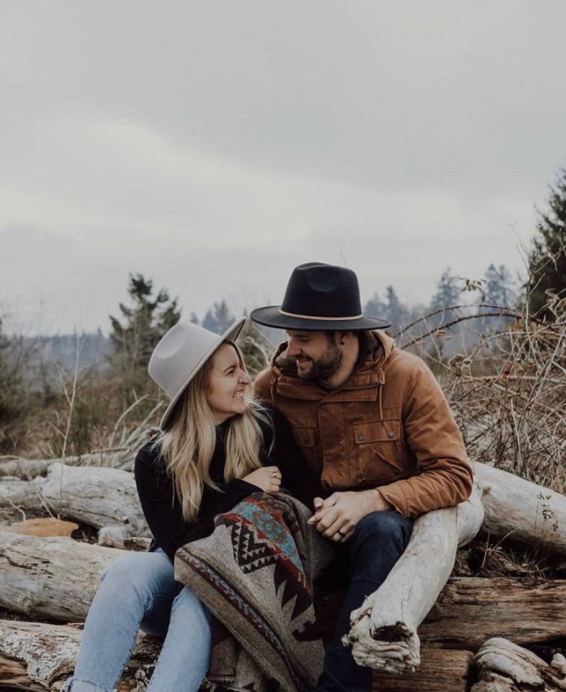 May include: A couple wearing hats and a blanket sit on a log in a wooded area. The woman is wearing a gray hat and a black sweater. The man is wearing a brown jacket and a black hat.