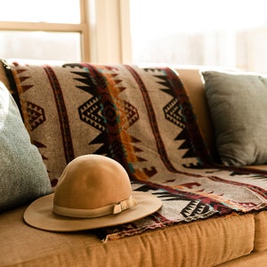 May include: A brown couch with a patterned throw blanket and two blue pillows. A brown fedora hat is resting on the couch.