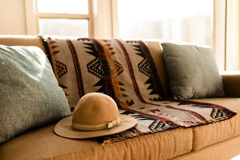 May include: A brown couch with a patterned throw blanket and two blue pillows. A brown fedora hat is resting on the couch.