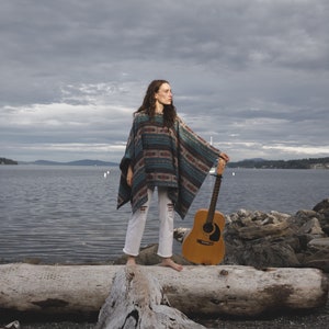 May include: A woman wearing a colorful poncho and white pants stands on a log holding an acoustic guitar. The background is a cloudy sky and a body of water.