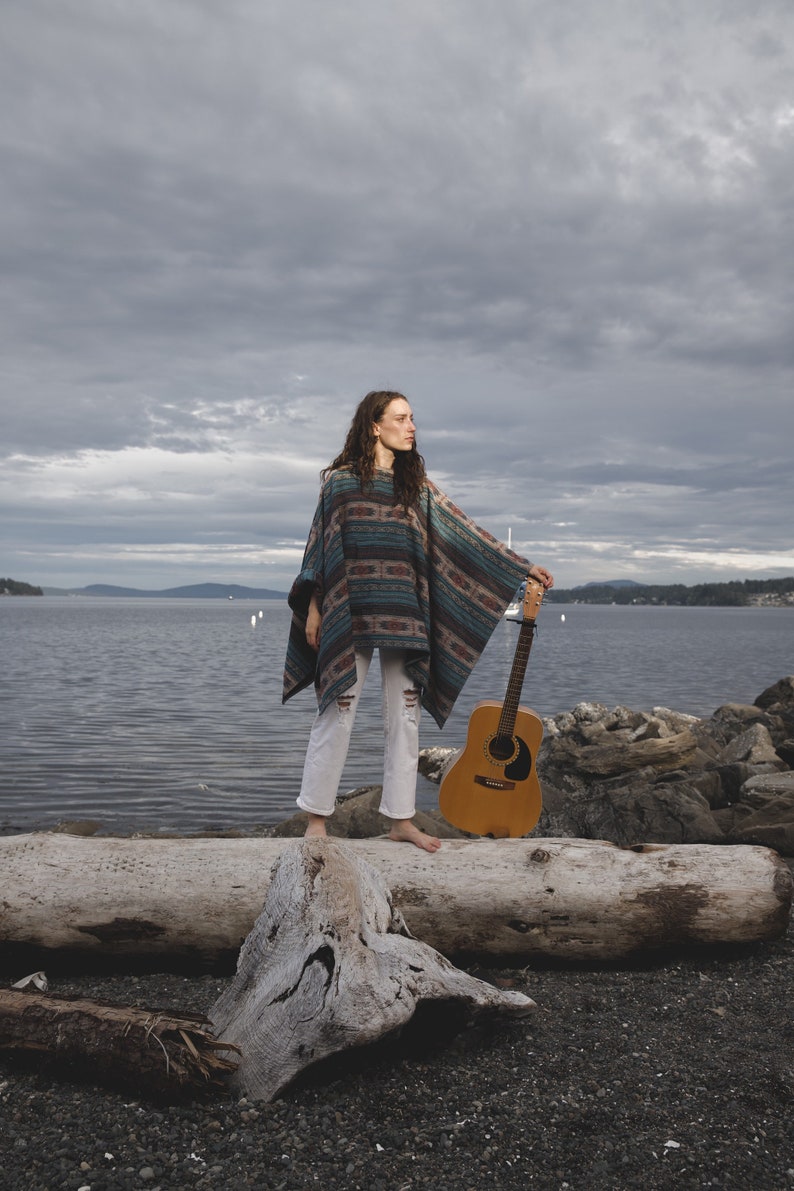 May include: A woman wearing a colorful poncho and white pants stands on a log holding an acoustic guitar. The background is a cloudy sky and a body of water.
