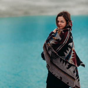 May include: A woman wearing a black shirt and black pants stands in front of a turquoise body of water. She is wrapped in a colorful patterned blanket with fringe.