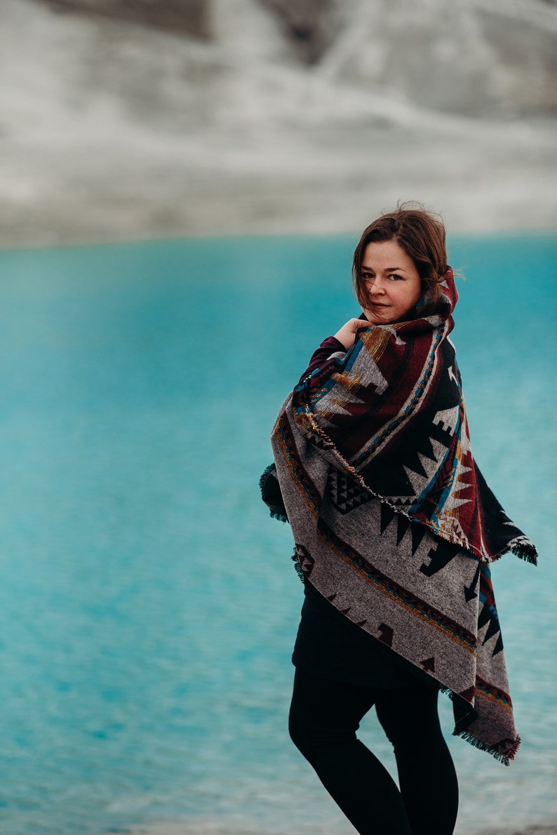 May include: A woman wearing a black shirt and black pants stands in front of a turquoise body of water. She is wrapped in a colorful patterned blanket with fringe.