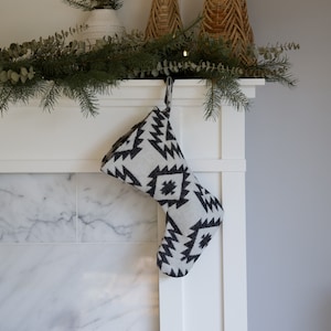 May include: A Christmas stocking with a black and white geometric pattern hangs from a white mantel. The stocking is made of a soft fabric and has a loop for hanging. The mantel is decorated with greenery and woven baskets.