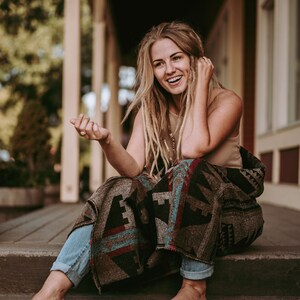 May include: A woman with long blonde dreadlocks sits on a set of stairs, wrapped in a colorful patterned blanket. She is smiling and looking to the side.