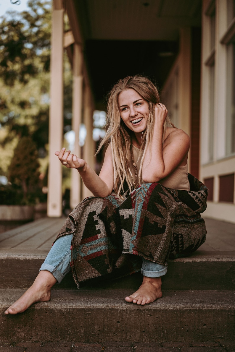 May include: A woman with long blonde dreadlocks sits on a set of stairs, wrapped in a colorful patterned blanket. She is smiling and looking to the side.
