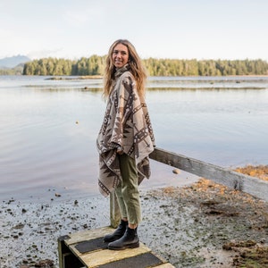 Puede incluir: Una mujer con un poncho estampado en marr&oacute;n y blanco y pantalones verdes se encuentra de pie en un muelle de madera sobre una masa de agua. Sonr&iacute;e y mira a la c&aacute;mara.