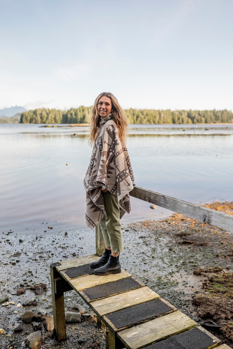 Puede incluir: Una mujer con un poncho estampado en marr&oacute;n y blanco y pantalones verdes se encuentra de pie en un muelle de madera sobre una masa de agua. Sonr&iacute;e y mira a la c&aacute;mara.