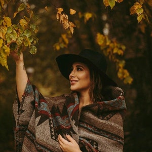 May include: A woman wearing a black hat and a brown and blue patterned poncho stands in a forest with fall foliage.