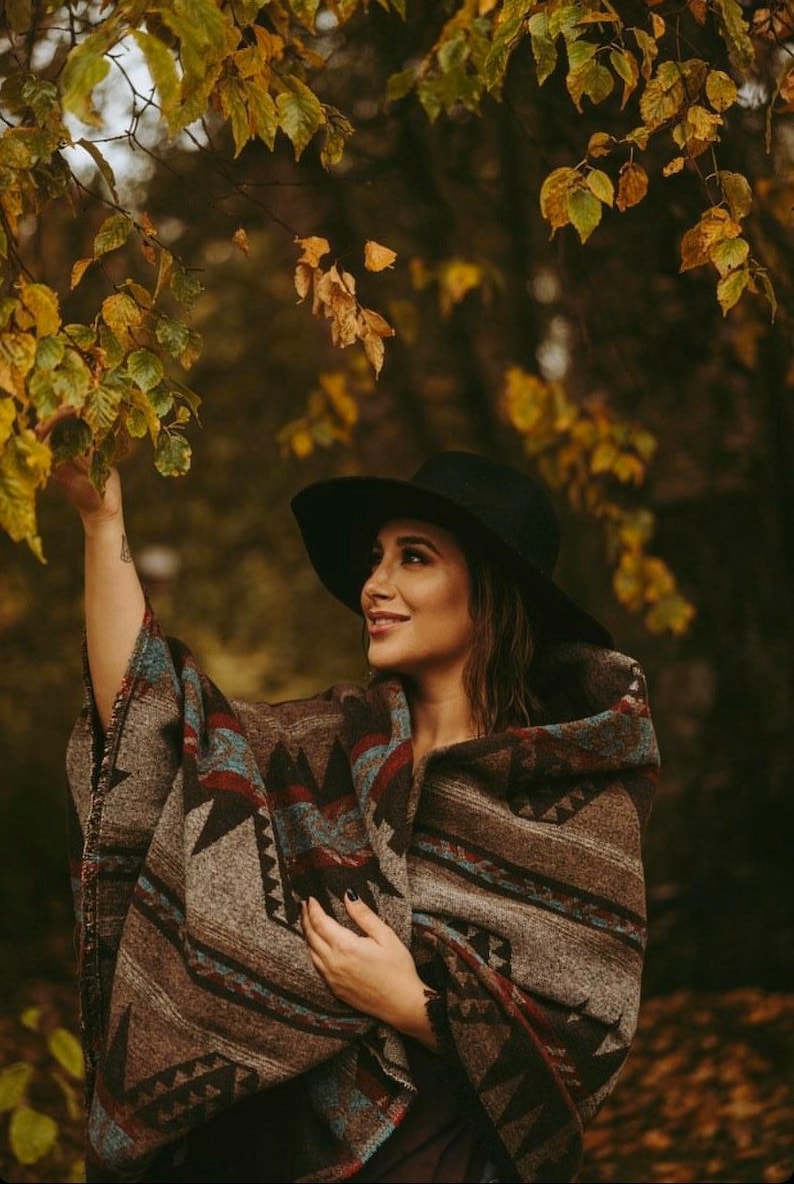 May include: A woman wearing a black hat and a brown and blue patterned poncho stands in a forest with fall foliage.