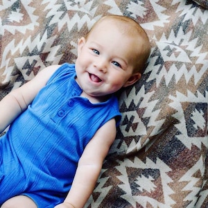 Könnte beinhalten: Ein kleiner Junge in einem blauen, ärmellosen Strampler lächelt, während er auf einer braunen und weißen gemusterten Decke liegt.