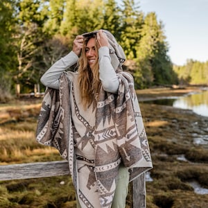 Puede incluir: Una mujer con un poncho con capucha con un patr&oacute;n geom&eacute;trico en tonos marrones y beige. Est&aacute; de pie junto a una valla de madera con un cuerpo de agua en el fondo.