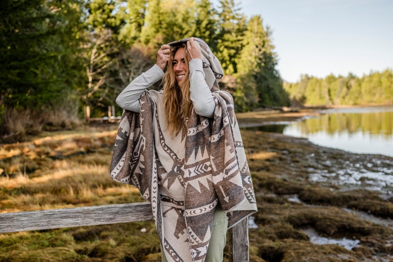 Puede incluir: Una mujer con un poncho con capucha con un patr&oacute;n geom&eacute;trico en tonos marrones y beige. Est&aacute; de pie junto a una valla de madera con un cuerpo de agua en el fondo.