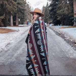 May include: A woman wearing a brown hat and a colorful patterned blanket stands on a snowy path in a forest.