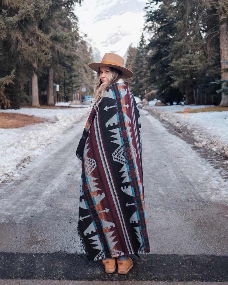 May include: A woman wearing a brown hat and a colorful patterned blanket stands on a snowy path in a forest.