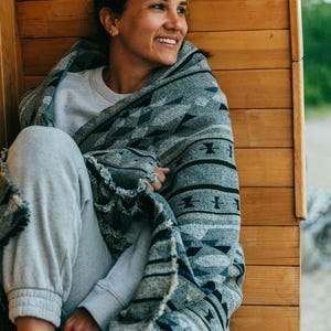 Puede incluir: Una persona envuelta en una manta gris y azul estampada, sentada en un banco de madera. La manta presenta diseños geométricos y flecos. La persona lleva una sudadera y un pantalón de chándal grises. El fondo es una pared de madera.