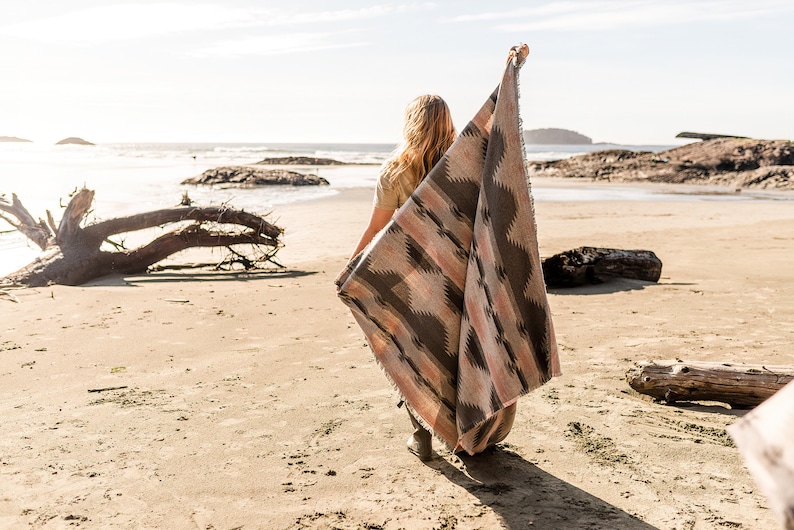 May include: A person standing on a sandy beach holding a woven blanket with a geometric pattern in shades of brown, pink, and black. The blanket is being held up in the air, and the person is looking out at the ocean.