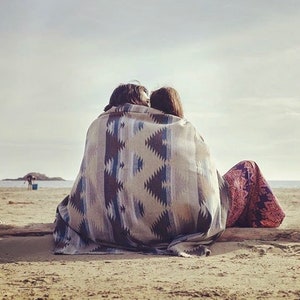 May include: A couple sits on a sandy beach, wrapped in a colorful blanket with a geometric pattern. They are facing away from the camera, looking out at the ocean.
