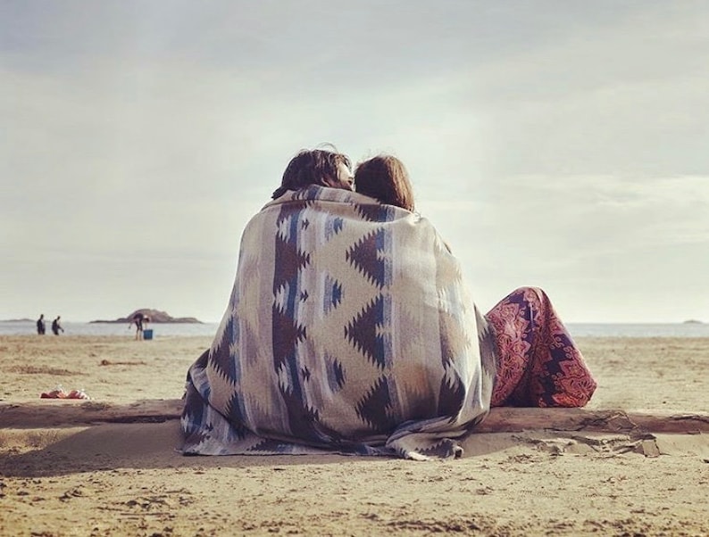 May include: A couple sits on a sandy beach, wrapped in a colorful blanket with a geometric pattern. They are facing away from the camera, looking out at the ocean.
