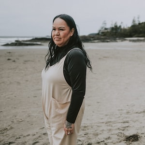 Puede incluir: Una mujer con un mono beige sobre una camisa de cuello alto negra se encuentra en una playa de arena. Tiene el pelo largo y negro y mira a la cámara.