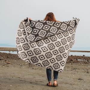 May include: A person standing on a beach holding a black and white patterned blanket over their head. The blanket has a geometric design.