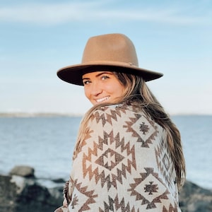 May include: A woman wearing a brown fedora and a white and brown patterned blanket. She is smiling and looking over her shoulder. The background is a blue sky and a body of water.