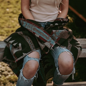May include: A person wearing ripped blue jeans and a brown, blue, and red patterned blanket wrapped around their waist. The person is sitting on a wooden bench.