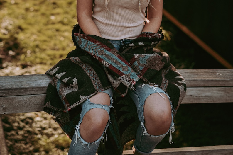 May include: A person wearing ripped blue jeans and a brown, blue, and red patterned blanket wrapped around their waist. The person is sitting on a wooden bench.