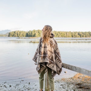 Puede incluir: Una persona con un poncho estampado en marr&oacute;n y blanco se encuentra de pie en un muelle de madera con vistas a una masa de agua. La persona lleva pantalones verdes y botas negras.