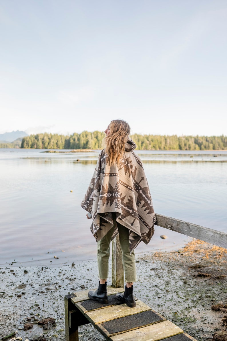 Puede incluir: Una persona con un poncho estampado en marr&oacute;n y blanco se encuentra de pie en un muelle de madera con vistas a una masa de agua. La persona lleva pantalones verdes y botas negras.