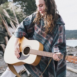 May include: A woman wearing a blue and brown patterned poncho plays an acoustic guitar with a brown strap. The guitar is a light brown color with a black pickguard.