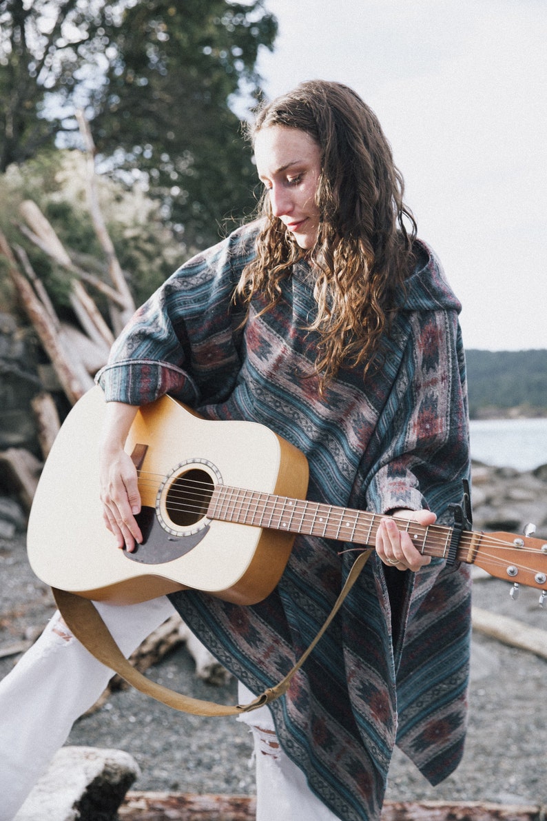 May include: A woman wearing a blue and brown patterned poncho plays an acoustic guitar with a brown strap. The guitar is a light brown color with a black pickguard.
