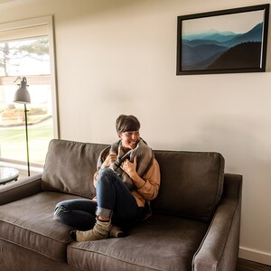 May include: A woman sits on a gray couch, wrapped in a gray and black blanket, smiling and looking to the right. The couch is in a living room with a large window and a framed print of a mountain range on the wall.