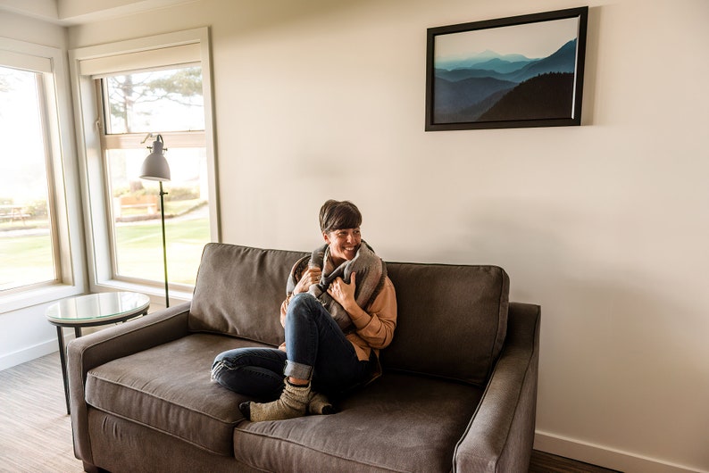 May include: A woman sits on a gray couch, wrapped in a gray and black blanket, smiling and looking to the right. The couch is in a living room with a large window and a framed print of a mountain range on the wall.