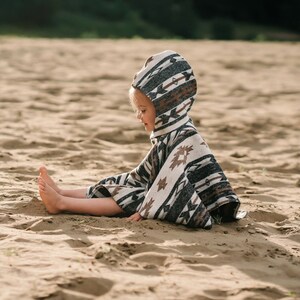 May include: A child wearing a hooded poncho with a geometric pattern sits on a sandy beach. The poncho is white, brown, and black with a geometric pattern.