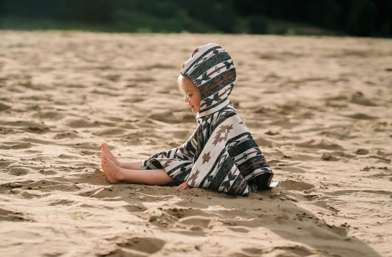 May include: A child wearing a hooded poncho with a geometric pattern sits on a sandy beach. The poncho is white, brown, and black with a geometric pattern.