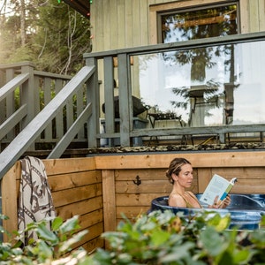 Puede incluir: Una mujer se relaja en una ba&ntilde;era de hidromasaje en una terraza de madera, leyendo un libro. La ba&ntilde;era de hidromasaje est&aacute; rodeada de paredes de madera y un arbusto verde. La terraza tiene una barandilla y escaleras que conducen a una caba&ntilde;a.