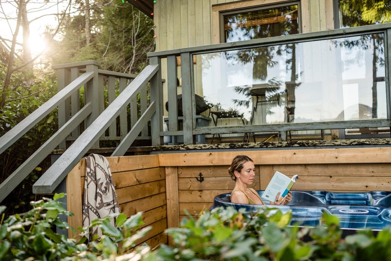 Puede incluir: Una mujer se relaja en una ba&ntilde;era de hidromasaje en una terraza de madera, leyendo un libro. La ba&ntilde;era de hidromasaje est&aacute; rodeada de paredes de madera y un arbusto verde. La terraza tiene una barandilla y escaleras que conducen a una caba&ntilde;a.