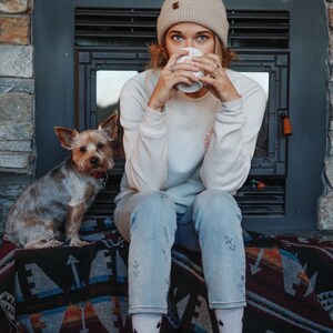 May include: A woman wearing a beige beanie and a white sweatshirt sits on a patterned blanket with a small dog. She is holding a white mug and looking down at it.