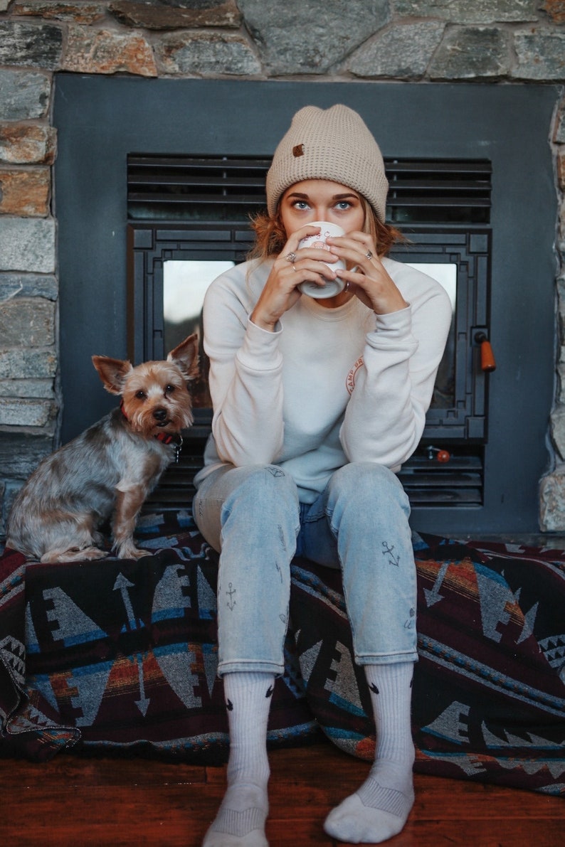 May include: A woman wearing a beige beanie and a white sweatshirt sits on a patterned blanket with a small dog. She is holding a white mug and looking down at it.