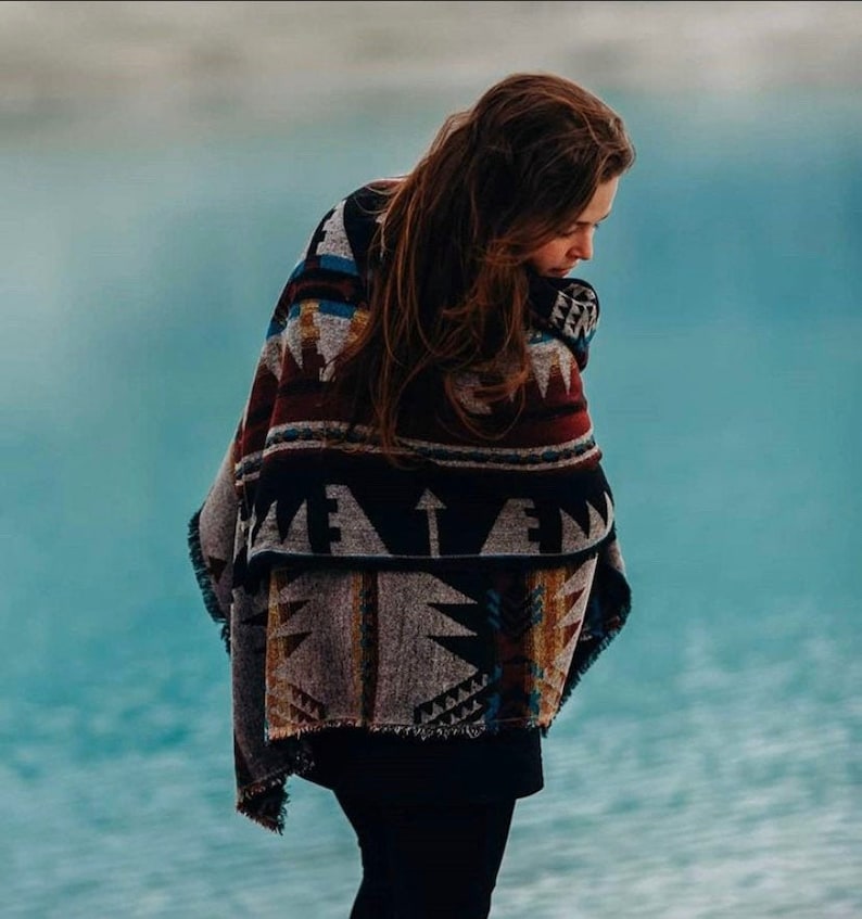 May include: A person wearing a colorful patterned blanket with black, white, red, and blue geometric designs. The blanket is wrapped around the person's shoulders and is fringed at the edges.