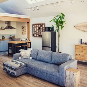 May include: A living room with a blue sectional sofa, a wooden coffee table, a potted plant, and a wooden shelf with a collection of items. The room has a light wood floor and white walls.