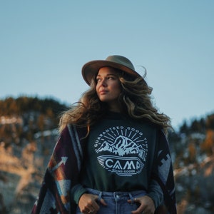 May include: A woman wearing a green long-sleeved shirt with a white graphic of a mountain range and the text "Guaranteed Good Times Camp Brand Goods" on the front. She is wearing a brown hat and a colorful patterned blanket over her shoulders. She is standing in front of a mountain range.