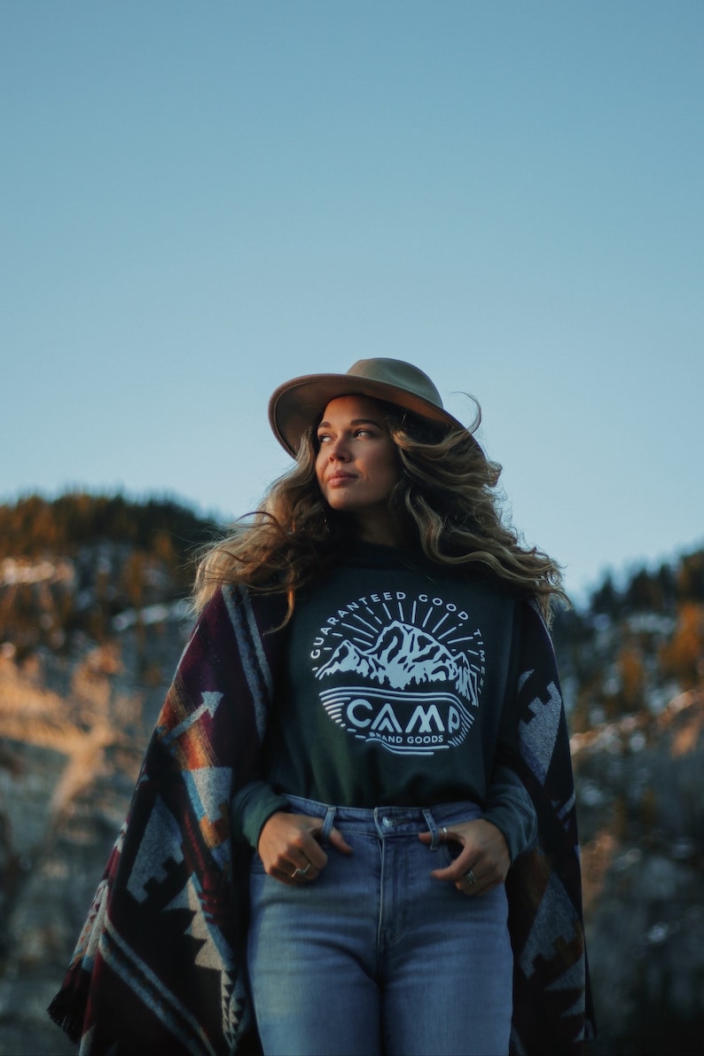 May include: A woman wearing a green long-sleeved shirt with a white graphic of a mountain range and the text "Guaranteed Good Times Camp Brand Goods" on the front. She is wearing a brown hat and a colorful patterned blanket over her shoulders. She is standing in front of a mountain range.