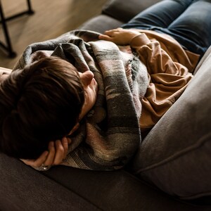 May include: A person is lying on a gray couch with a brown and gray blanket. They are wearing a brown shirt and blue jeans.